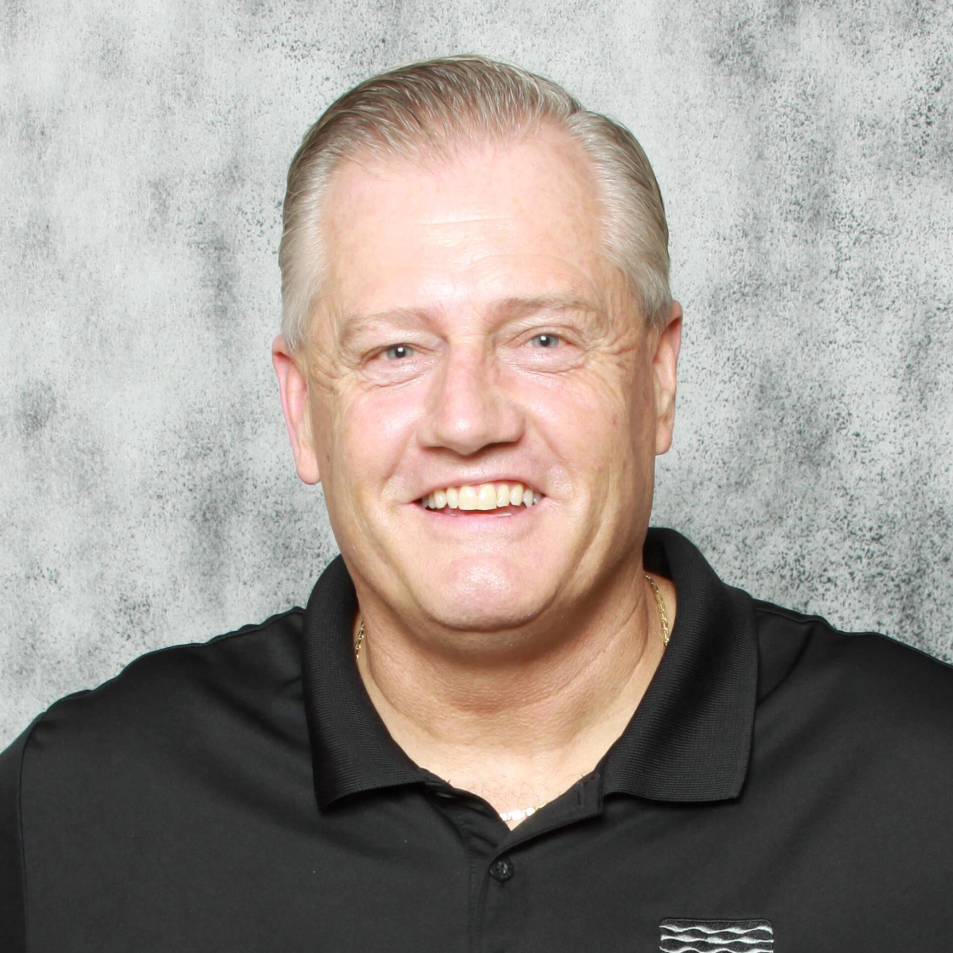 A smiling person with short, light-colored hair, wearing a black collared shirt, against a mottled gray background. Appears happy and approachable.