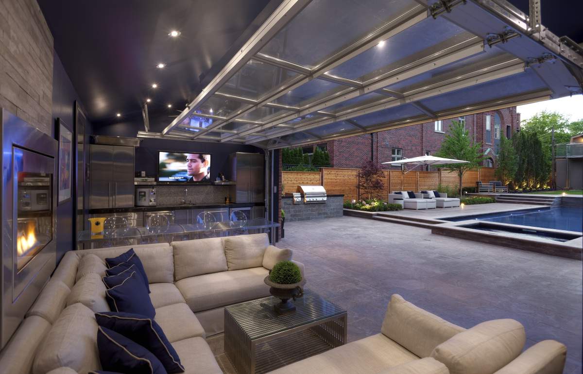 Luxurious outdoor living space with a sectional sofa, fireplace, TV, kitchen area, and a view of a pool with lounging chairs in the background.