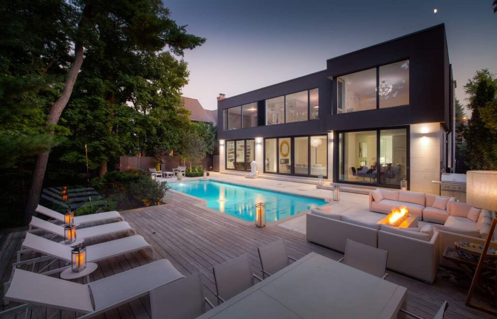 A modern two-story house with large windows lit from inside, a pool, deck chairs, outdoor couches, and a fire pit under twilight skies.