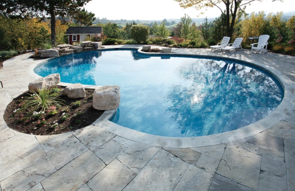 A serene backyard setting with a large, kidney-shaped swimming pool surrounded by stone paving and lounge chairs, overlooking a lush landscape.