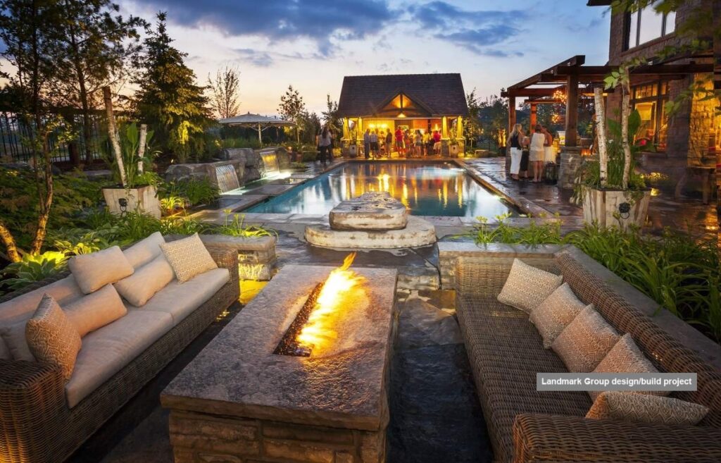 An outdoor luxury living space with a pool, fire feature, comfortable seating, lush landscaping, and a group of people gathering at dusk.