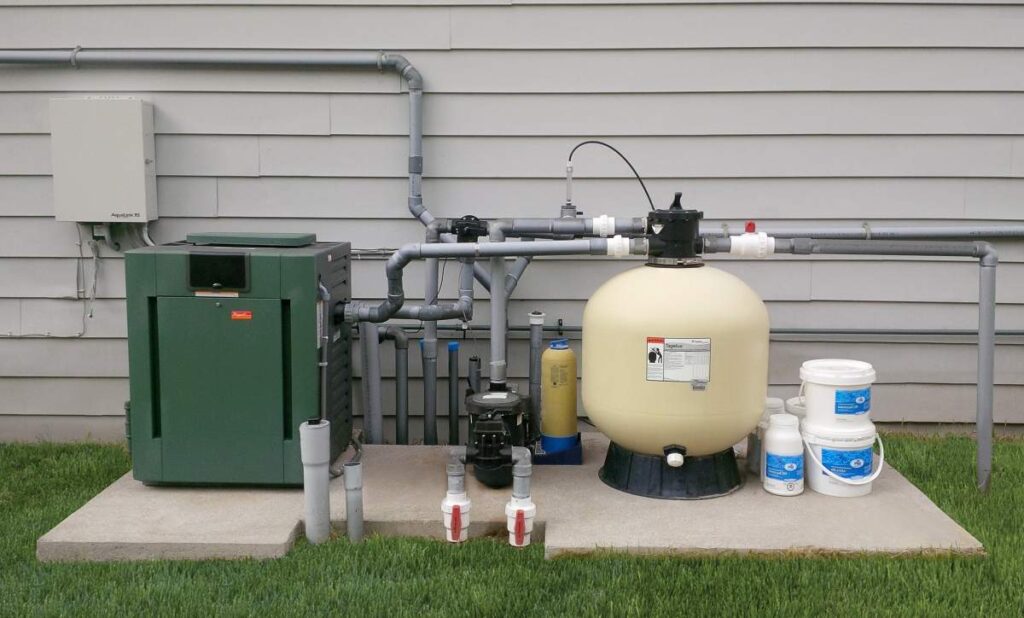 Outdoor pool equipment setup with a filtration system, pump, heater, and chemical containers arranged neatly on a concrete pad against a house siding.