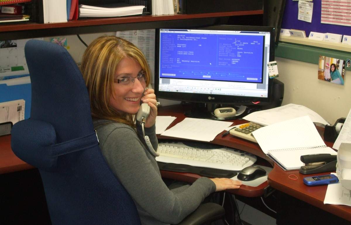 A person is seated at an office desk, using a phone, with a computer displaying blue screens, papers, and personal items surrounding them.
