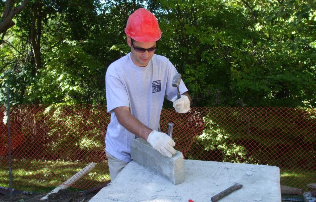 A person wearing a red hard hat and safety glasses is carving stone with a chisel and hammer, standing by a construction barrier outside.