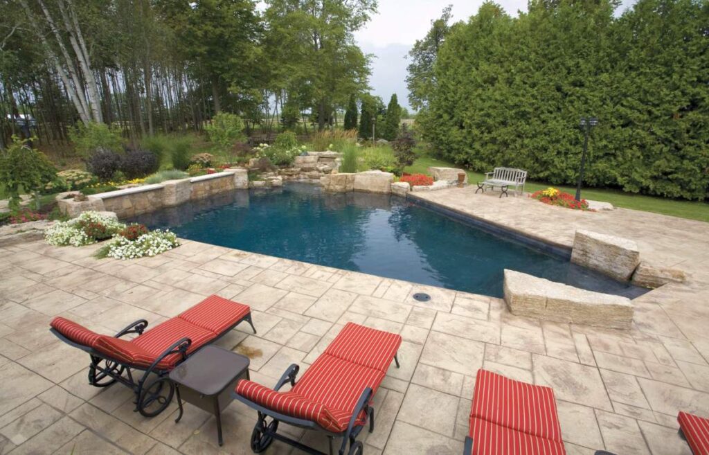 An outdoor pool surrounded by a stone patio with loungers, landscaping, and trees under a partly cloudy sky, suggesting a peaceful, private backyard oasis.