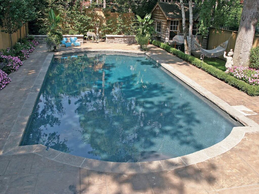 A serene backyard with a rectangular swimming pool, stone patio, lush plants, a hammock, wooden playhouse, sculptures, and colorful flower beds under a clear sky.