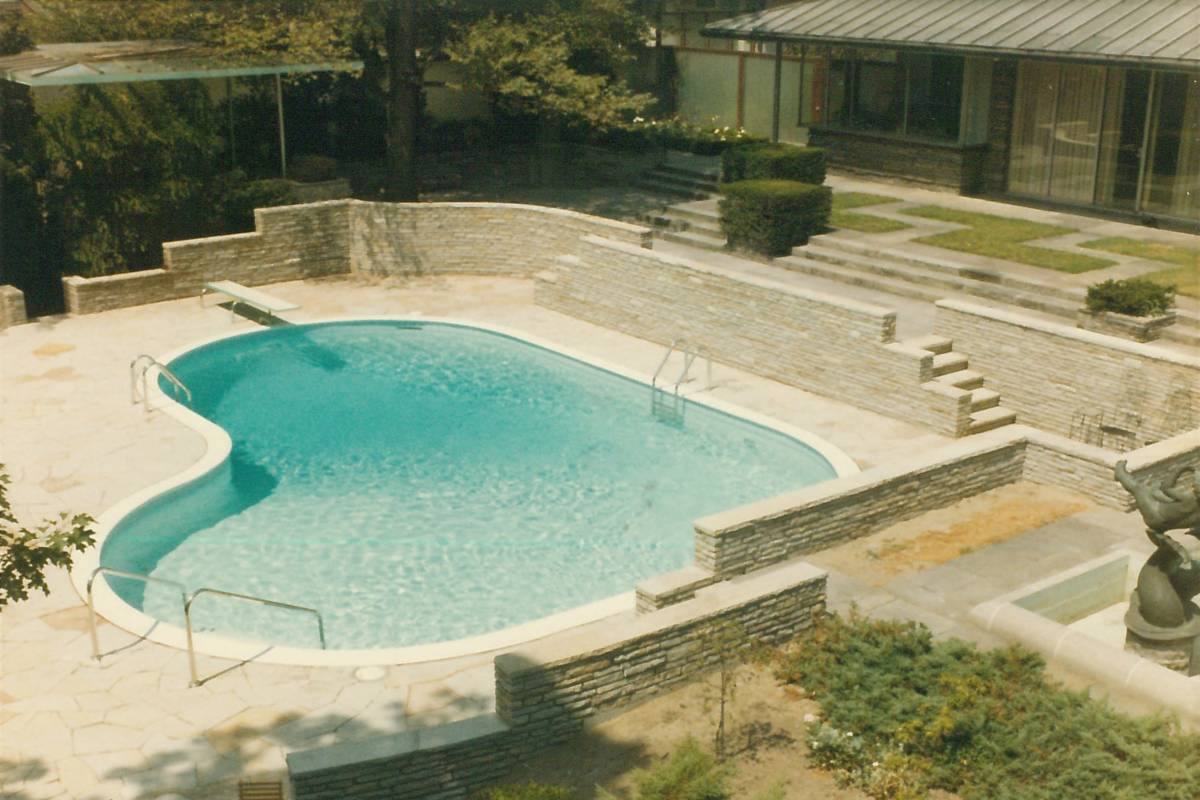 A kidney-shaped swimming pool is surrounded by stone walls and steps, with landscaped grass and shrubs adjacent to a modern building. No people visible.