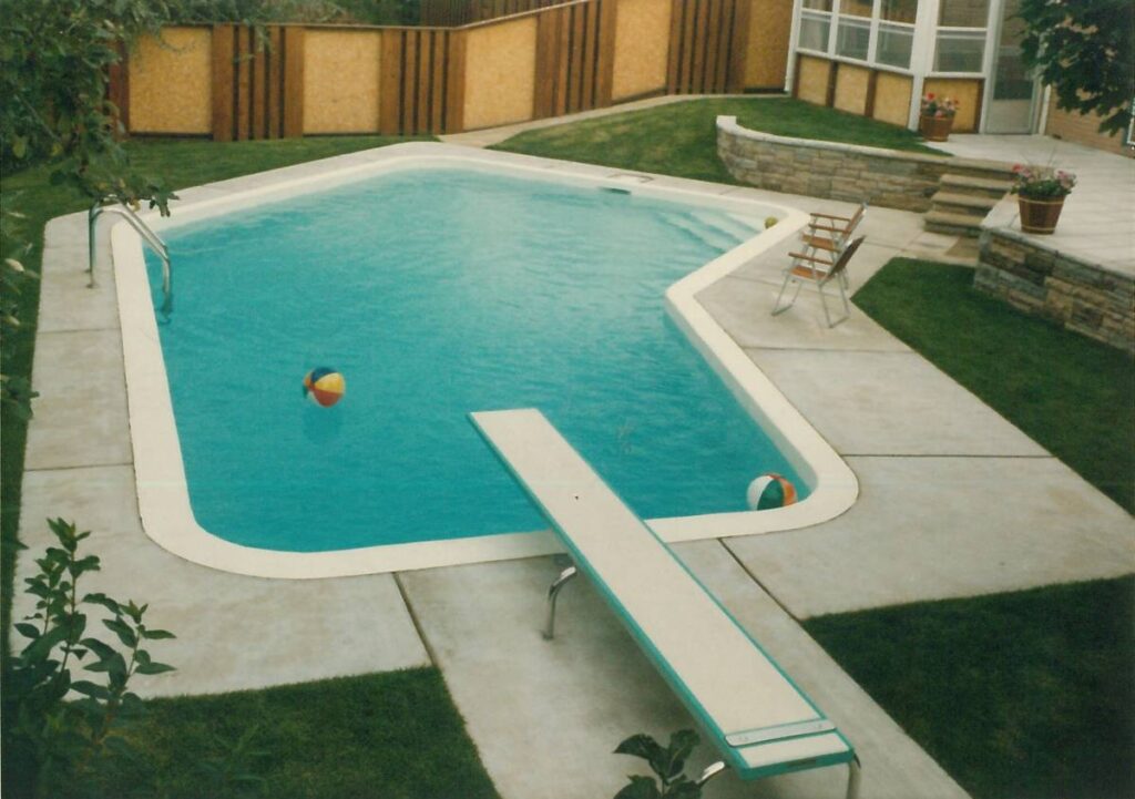A residential backyard with a blue in-ground swimming pool, concrete walkway, diving board, green lawn, fence, and a folding chair nearby.