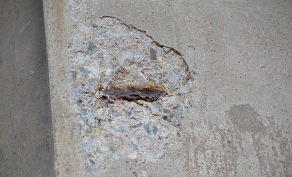 The image shows a close-up of a deteriorated concrete surface with exposed, rusted steel reinforcement suggesting structural damage or aging.