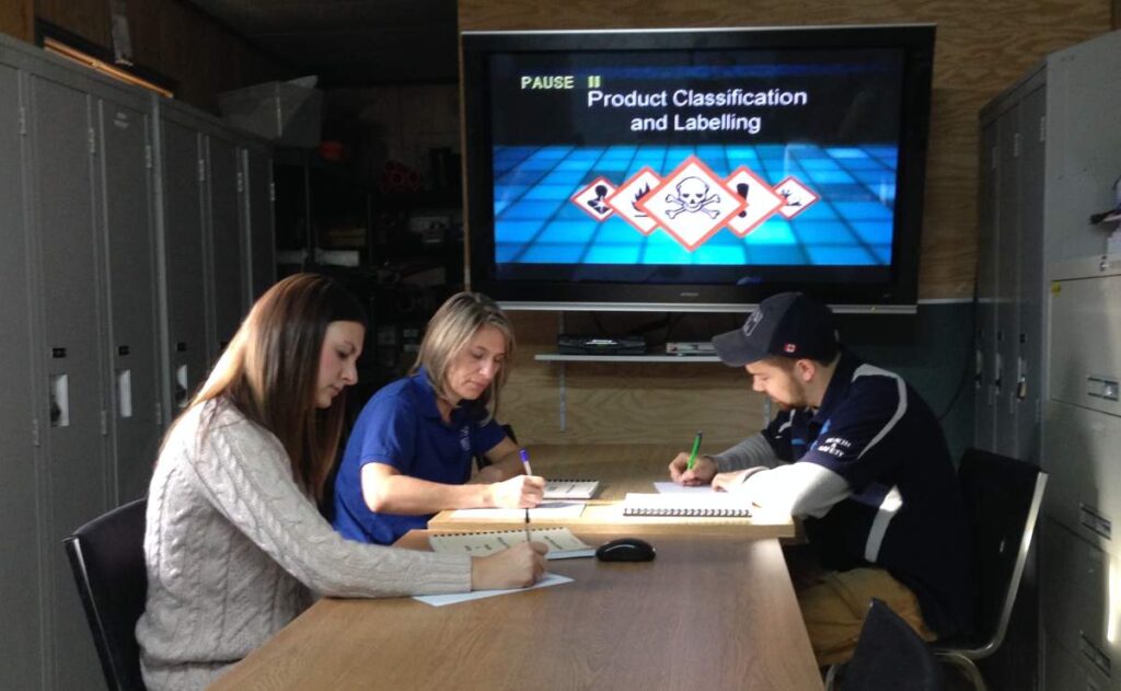 Three people sit at a table writing notes, with a presentation titled "Product Classification and Labelling" displayed on a screen behind them.