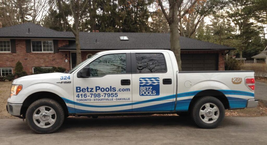 A white and blue pickup truck with "Betz Pools" branding is parked in front of a brick house with a treed driveway.