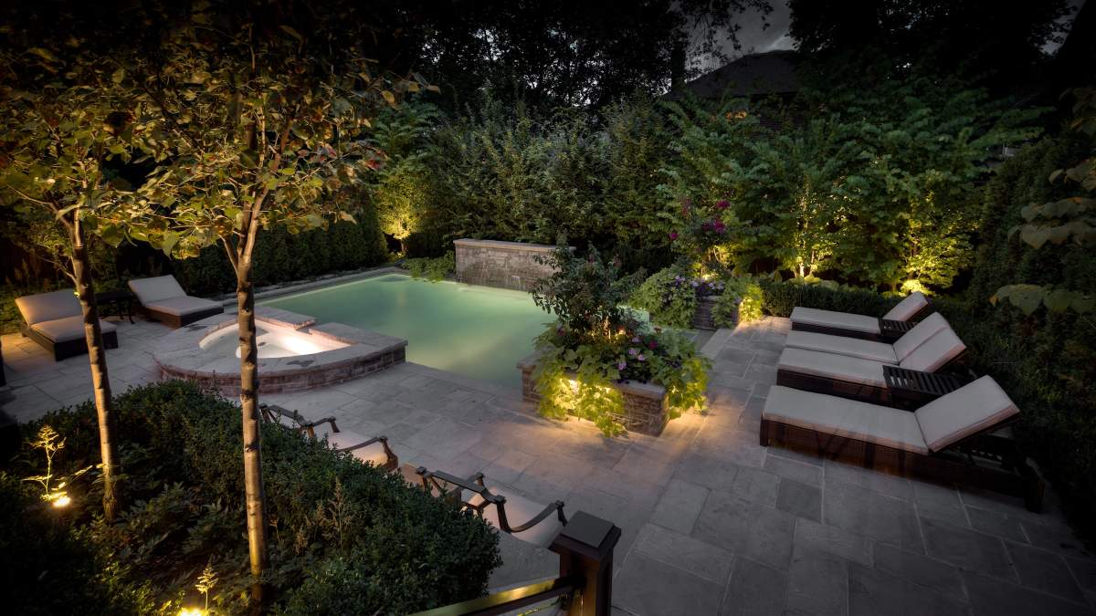 This image features an elegant outdoor space at dusk with a swimming pool, surrounded by lush greenery and trees, and multiple lounge chairs for relaxation.