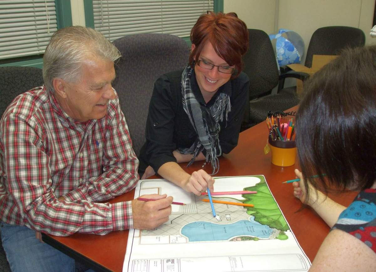 Three people are seated around a table, focused on coloring a large illustration with colored pencils, smiling, in a room with a globe and blinds.