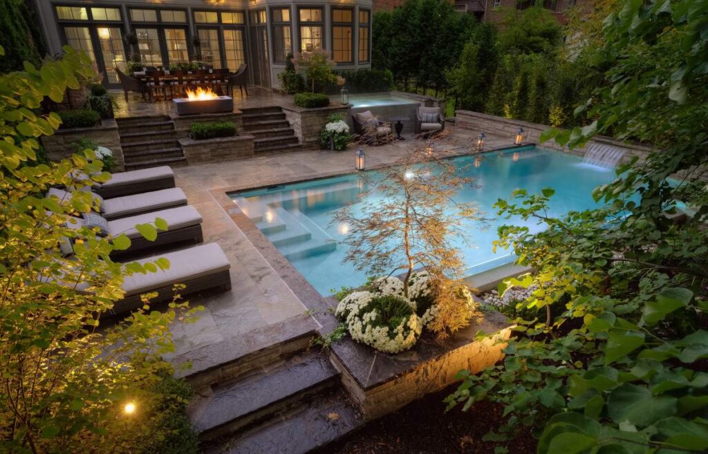 A luxurious backyard at dusk featuring an illuminated swimming pool, stone steps, sun loungers, a fire pit, landscaped plants, and an adjoining house.