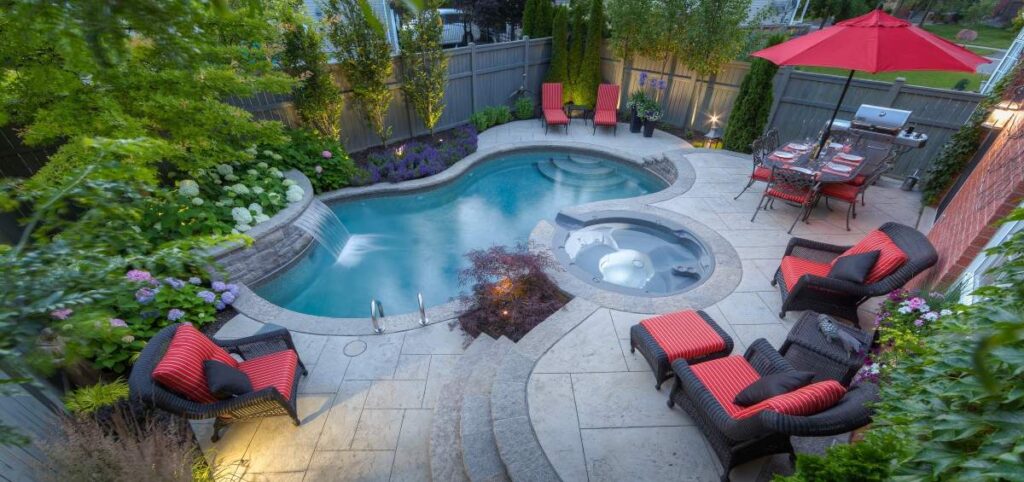 An inviting backyard with a curvy swimming pool, hot tub, outdoor furniture, a BBQ grill, and a red umbrella during dusk. Greenery surrounds the tranquil area.