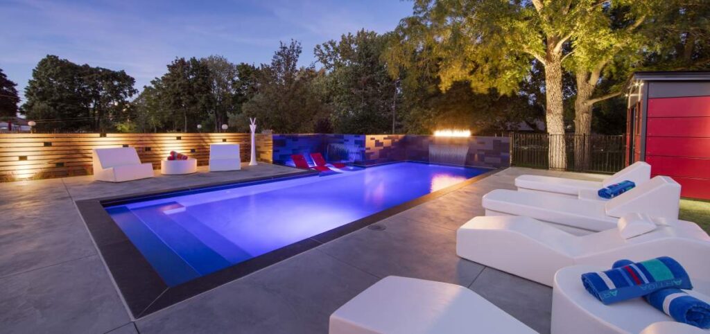 An outdoor swimming pool illuminated at dusk with modern lounge furniture, a red shed, ambient lighting, and surrounding trees in a backyard setting.