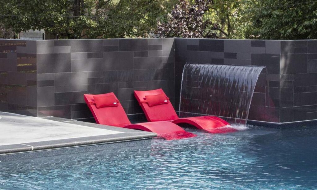A serene swimming pool with a dark tile surround, featuring a sleek waterfall and two vibrant red lounge chairs partially submerged in water.