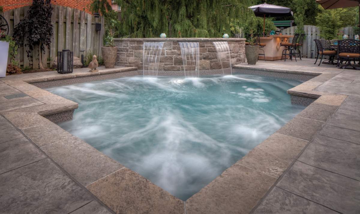 An outdoor residential setting featuring a square spa with cascading waterfalls, surrounded by paving stones, with patio furniture and greenery in the background.