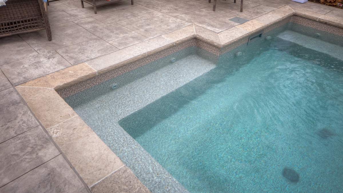 The image shows a modern swimming pool with clear blue water, surrounded by a tiled deck, with a bench, and steps visible on one side.