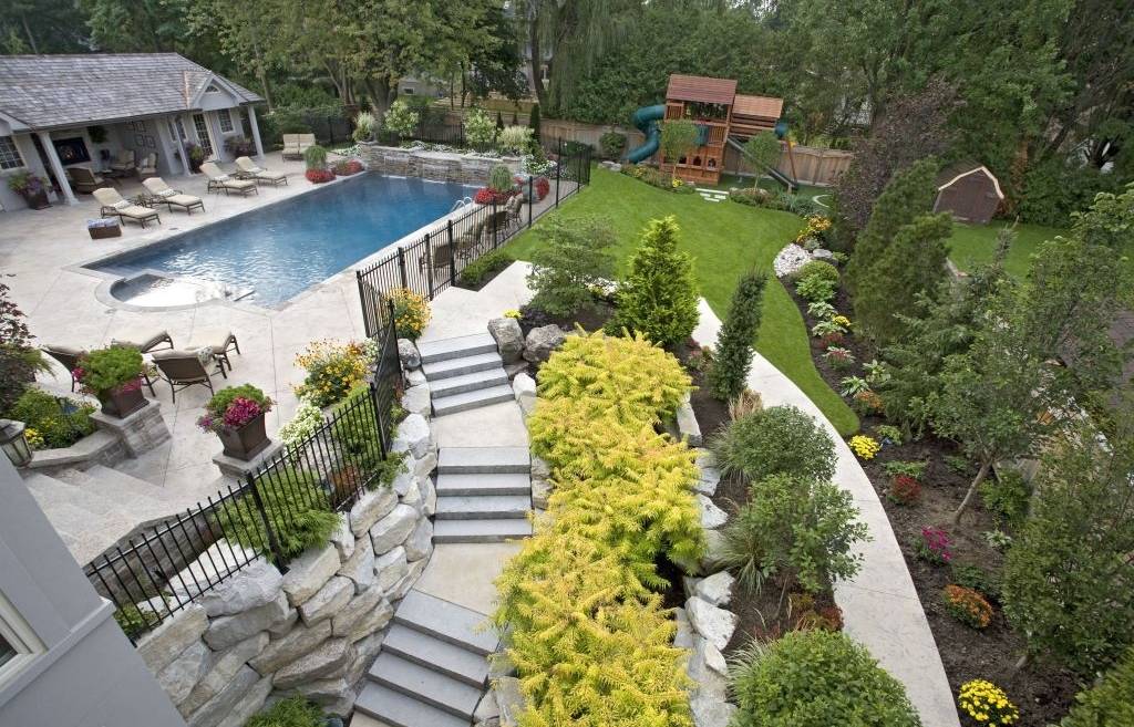 This is a well-manicured backyard with a swimming pool, lounge chairs, colorful flowerbeds, a play structure, lawn, and a stone stairway amid lush greenery.