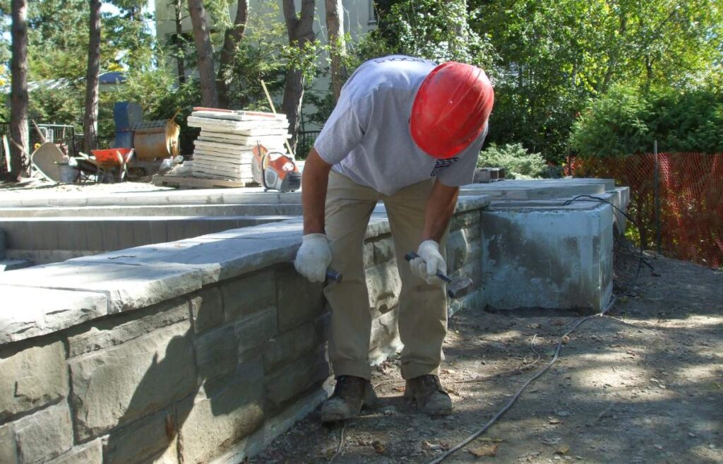 A person wearing a hard hat and gloves is working on a stone wall with a hammer and chisel, with construction materials in the background.