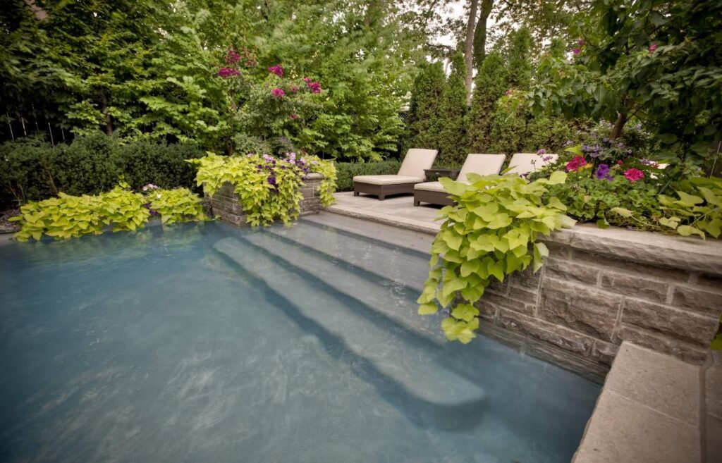 An outdoor swimming pool with clear blue water flanked by a stone wall, sun loungers, and vibrant, flowering plants under a verdant treeline.