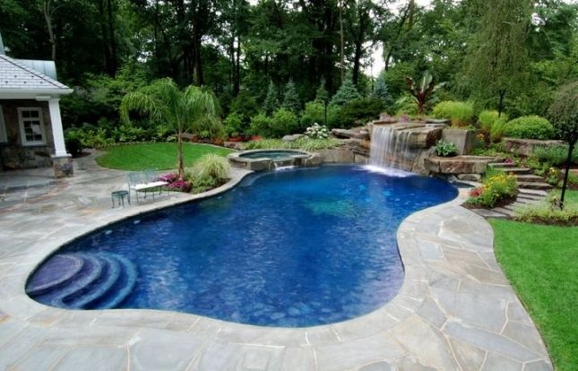 A luxurious backyard with a curvilinear inground swimming pool featuring a waterfall, surrounded by a stone patio, lush landscaping, and a small outbuilding.