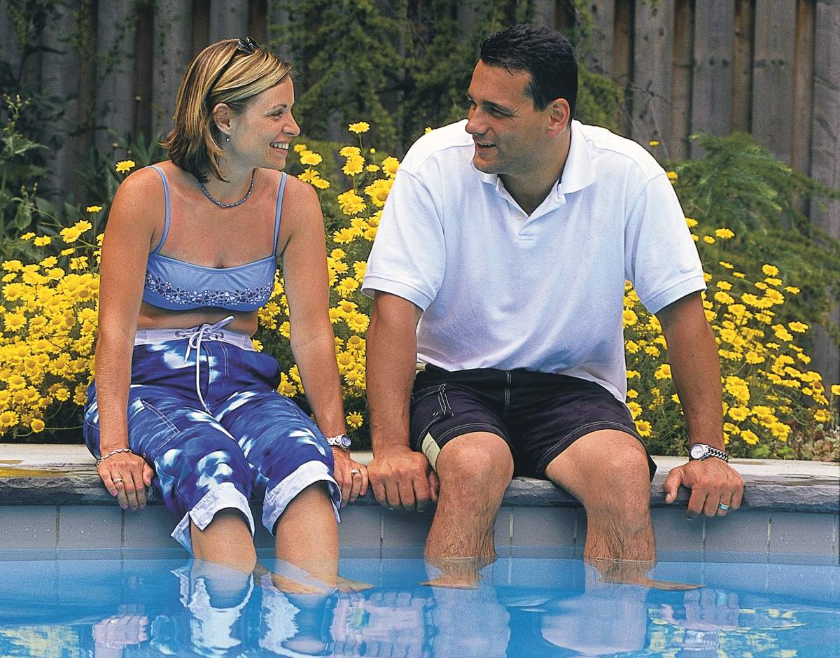 Two people sitting by a pool with their feet in the water, smiling, surrounded by yellow flowers and greenery, wearing casual summer clothing.