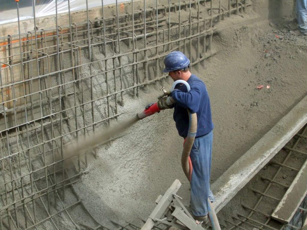 A person in a hard hat and safety gear is spraying concrete on a wall at a construction site, a technique known as shotcreting.