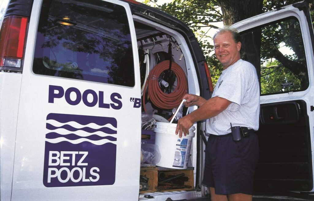 A person stands beside a white van labeled with "Betz Pools," holding a bucket and hose, smiling at the camera, with trees in the background.