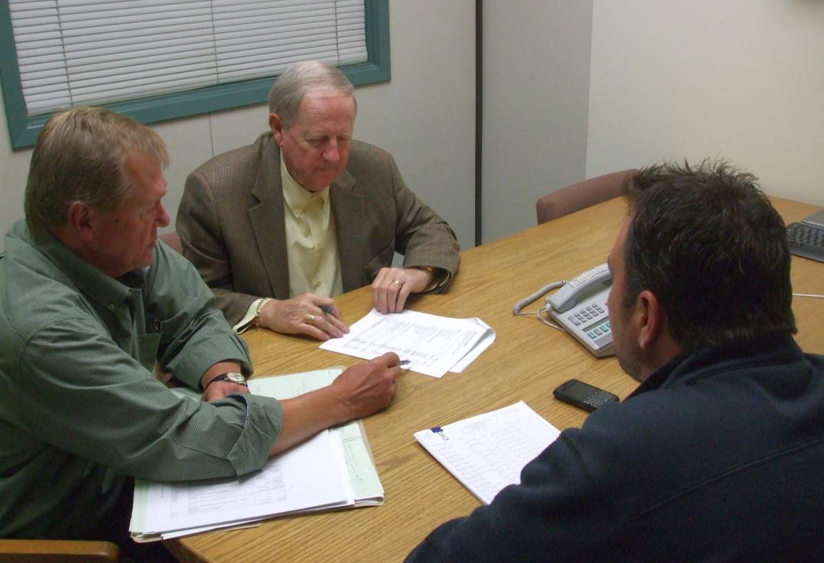 Three people are engaged in a meeting around a table, reviewing documents, with a phone and notepad visible, in a room with a window.