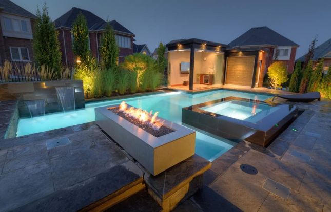 A luxurious backyard at dusk with illuminated pools, a fire feature, landscaped garden, modern design elements, and a cozy ambiance. No people visible.