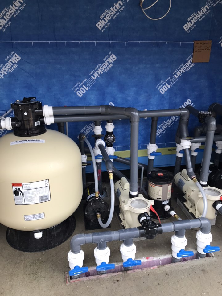 This image shows a complex arrangement of pool equipment including pipes, a sand filter, pumps, and valves against a blue waterproofing membrane background.