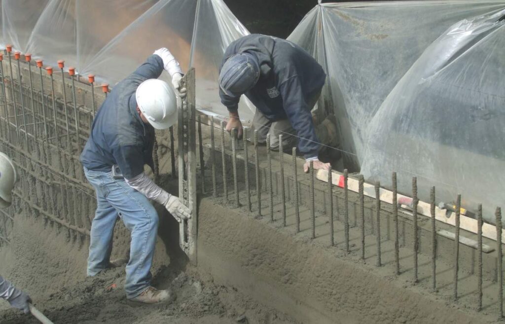 Two persons in hard hats and gloves work with wet concrete and rebar at a construction site, with a tarp protecting the area.