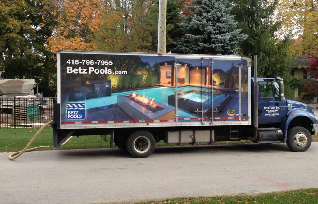 A truck with a pool company advertisement on its side is parked on a suburban road, featuring images of luxurious pools and contact information.