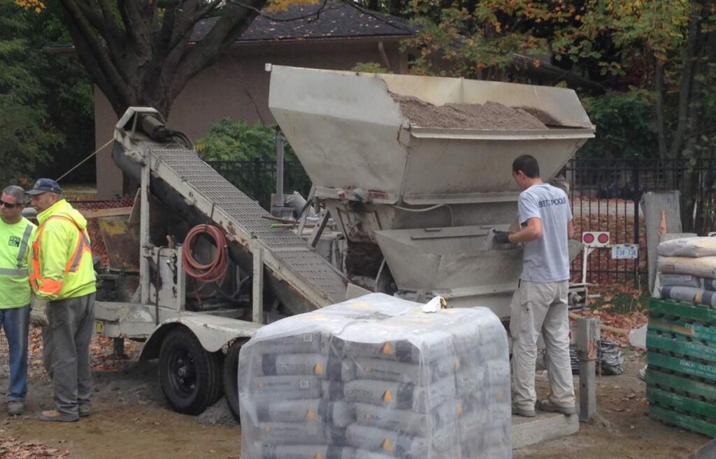 A construction site with three people, a cement mixer truck, and construction materials. One person is operating the mixer, another is observing.
