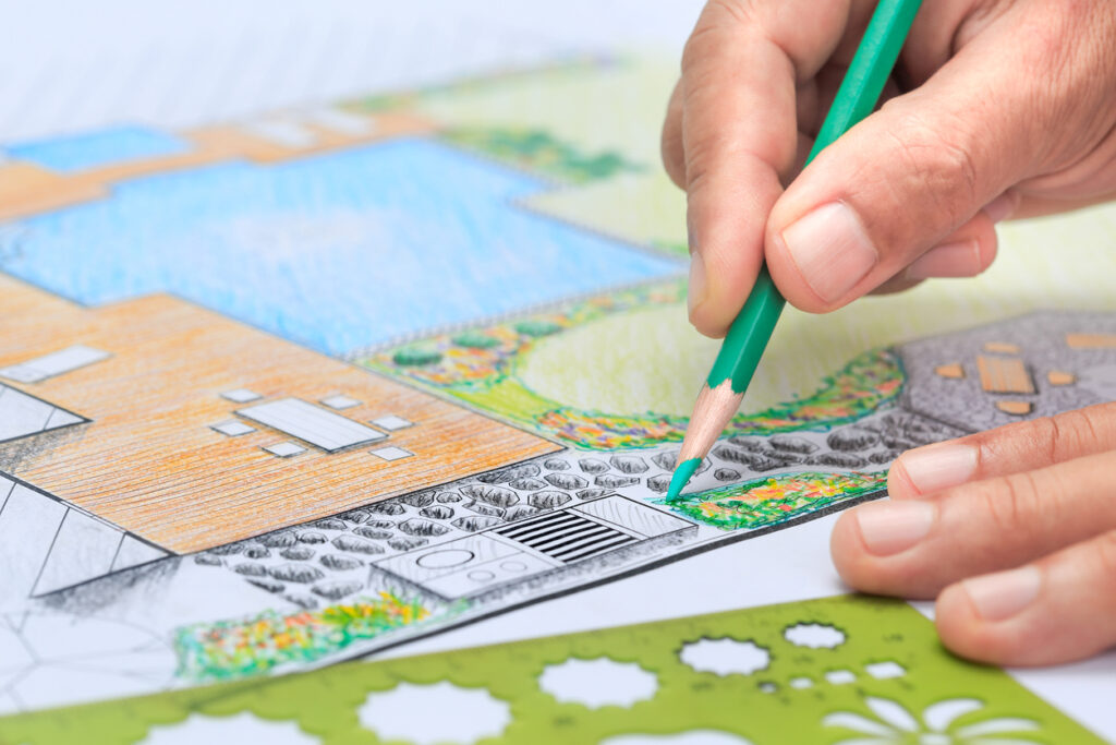 A person's hand is coloring a detailed landscape architecture plan with a green pencil, adding textures to trees and grass areas.