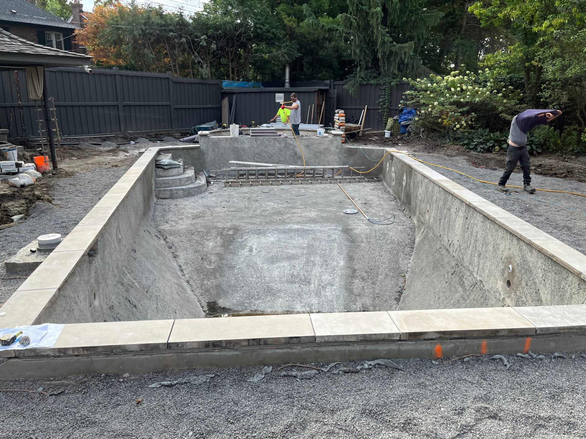 An unfinished swimming pool with two people working on the site. Construction materials are visible, and the pool is set in a backyard.