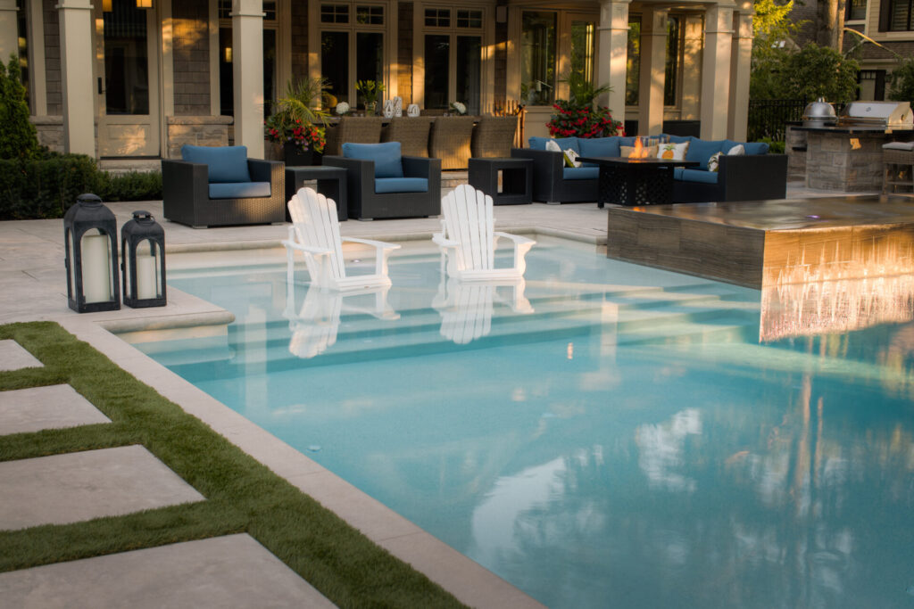 An elegant backyard with a swimming pool, two white chairs in the water, surrounded by a seating area, outdoor fireplace, and landscaping at dusk.