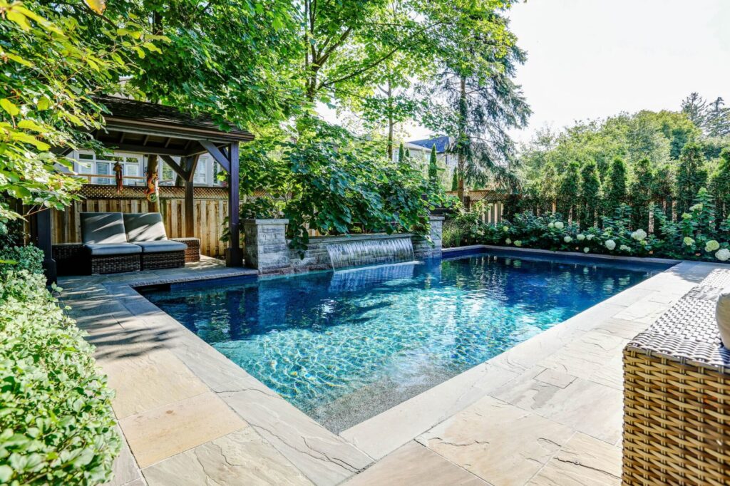 An outdoor swimming pool with clear blue water, surrounded by lush greenery. A shaded lounging area and elegant stone patio enhance the tranquil setting.