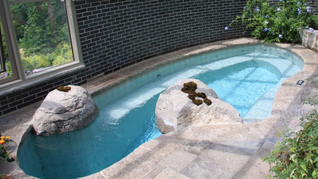 An outdoor kidney-shaped swimming pool with clear blue water, nestled beside a black brick house with large windows and surrounded by a stone patio.