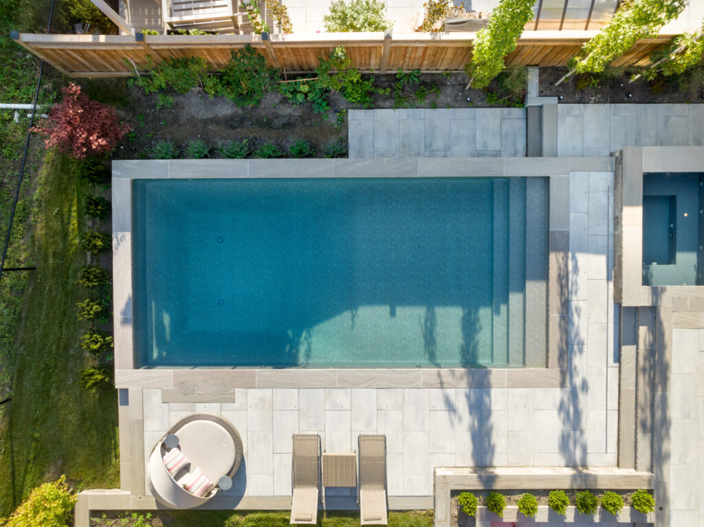 An aerial view of a backyard with a large rectangular swimming pool, sun loungers, circular seating area, landscaped greenery, and concrete paving.