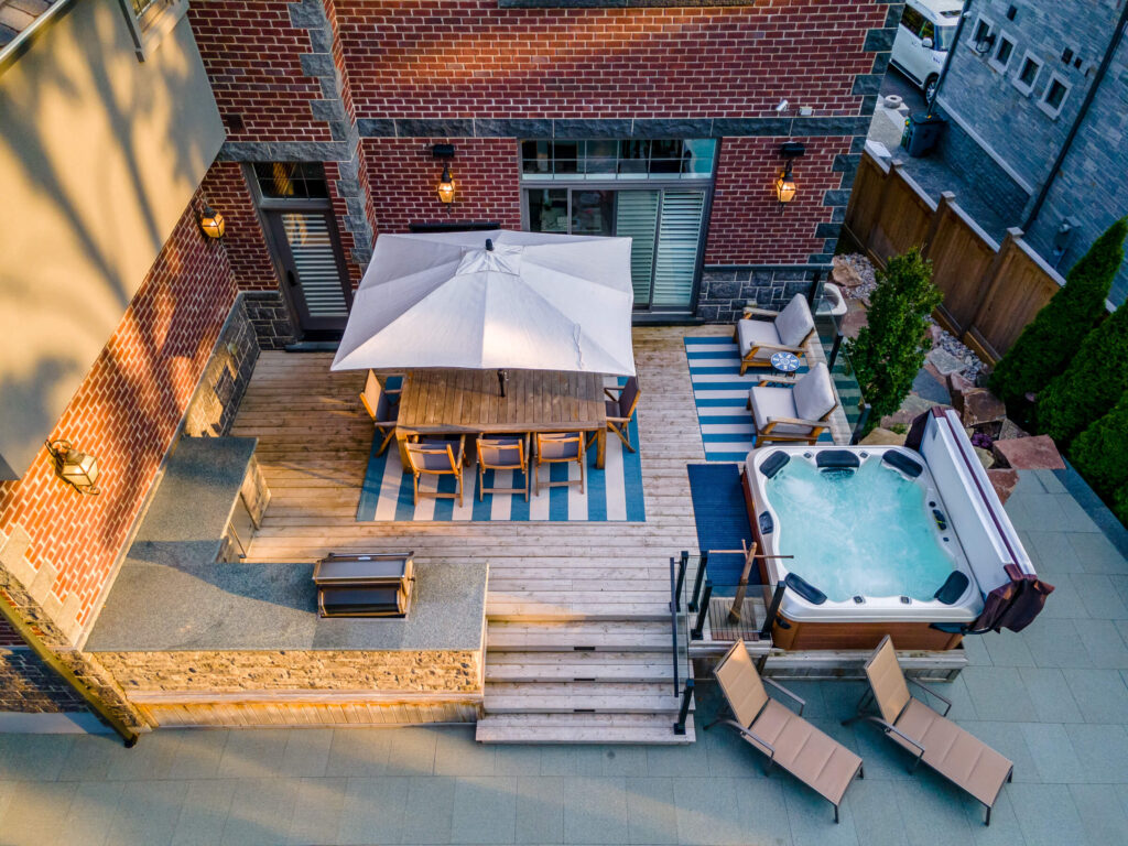 An aerial view of a backyard with a dining area, lounge chairs, a hot tub, a pool, and landscaping by a brick house during twilight.