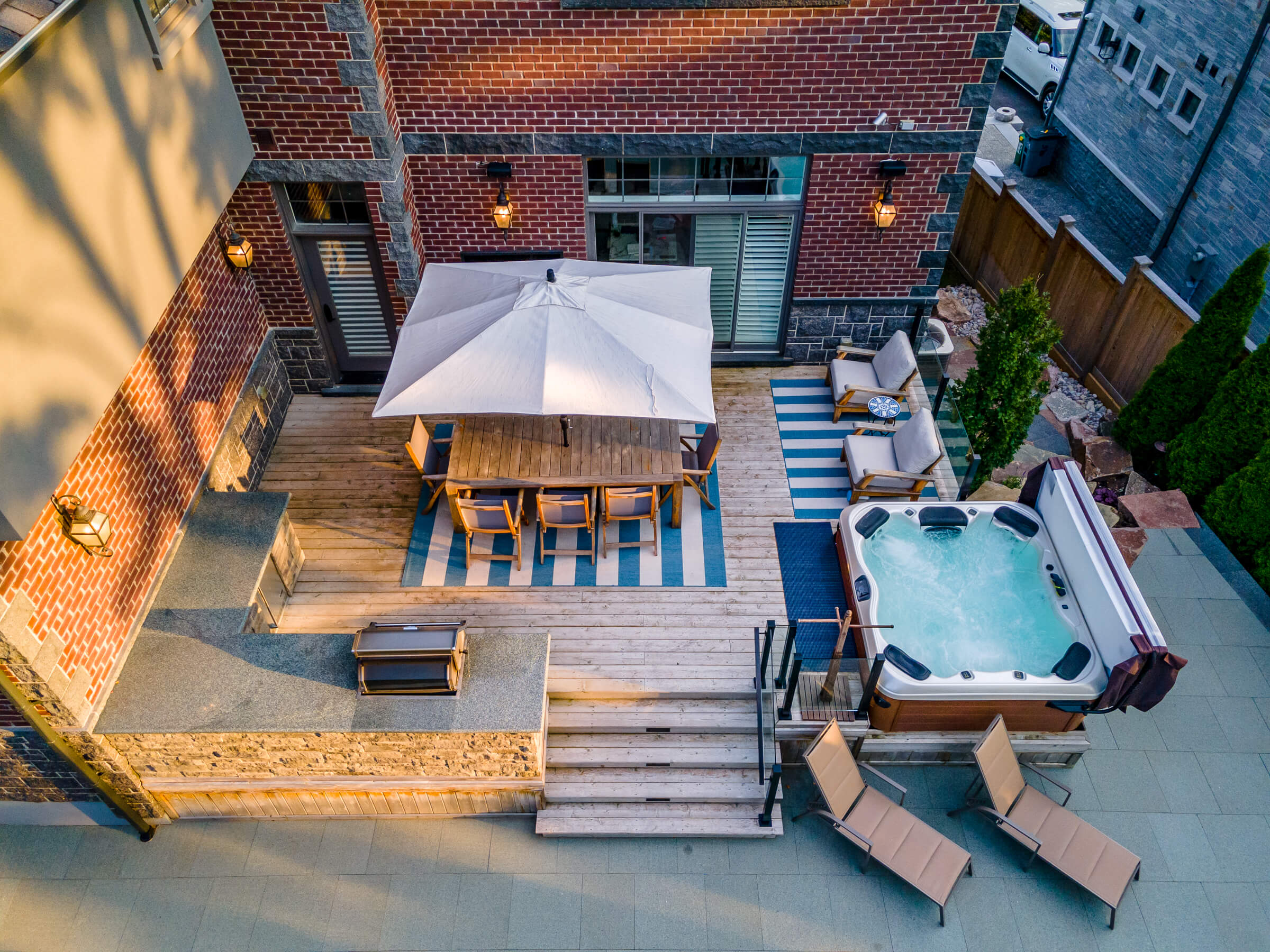 An aerial view of a backyard with a dining area, lounge chairs, a hot tub, a pool, and landscaping by a brick house during twilight.