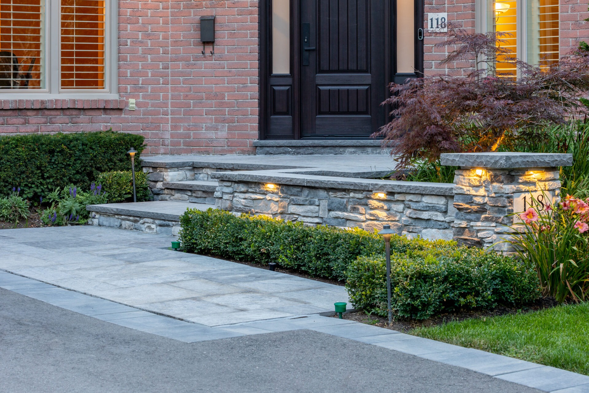 A well-manicured front yard with brick walls, landscaped garden, stone steps, lit with outdoor lighting, leading to a dark wooden door and brick house.