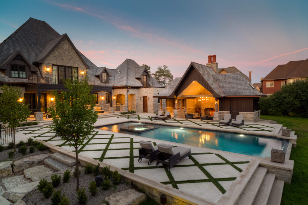 Luxurious house with stone façade, steep roofs, an outdoor swimming pool, and a patio area with a fireplace during a sunset with pinkish skies.