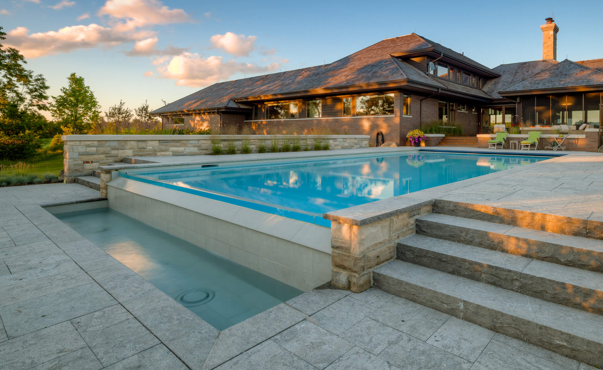 An elegant backyard with a clear blue swimming pool in the evening light, featuring steps, a patio, and a luxurious house with large windows.