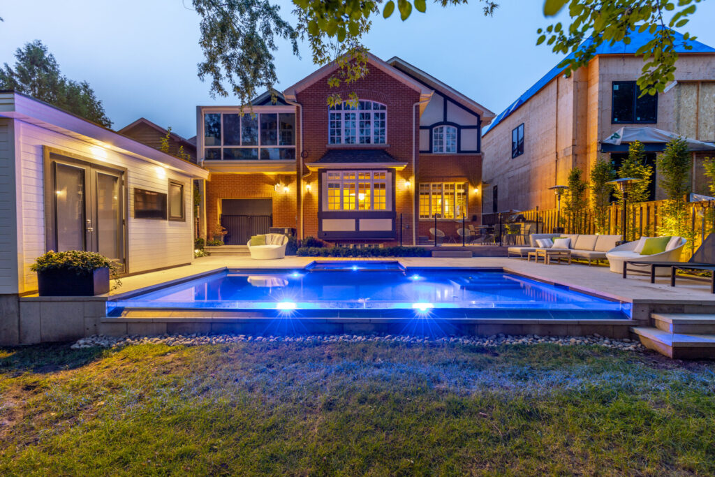 A luxurious evening setting with an illuminated swimming pool, deck chairs, a well-lit house, and an outbuilding, showcasing upscale outdoor residential architecture and landscaping.
