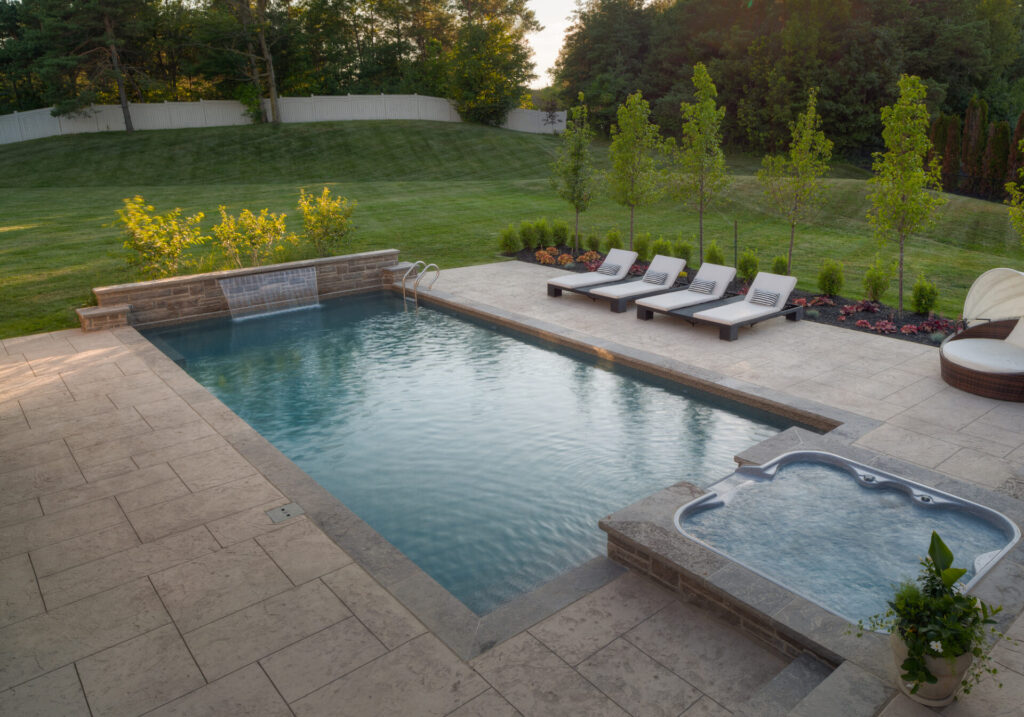 An outdoor swimming pool with an adjacent hot tub, surrounded by lounge chairs, and a well-maintained garden with trees during the evening.