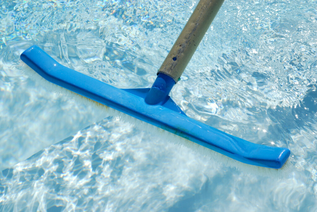 This image features a blue pool brush with a wooden handle submerged in clear, sparkling swimming pool water, reflecting sunlight on a bright day.